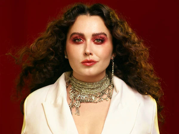 Portrait of an adult woman with curly hair, wearing a white blazer and a jeweled collar, against a red studio backdrop.