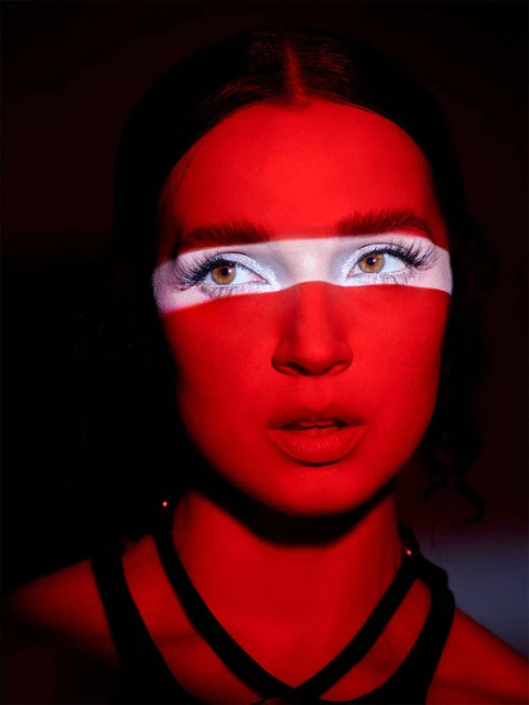 Close-up portrait of a person with striking red and white face paint and black strapped clothing, set against a dark, shadowy background.