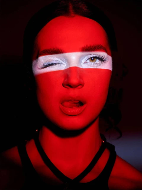 Portrait of a woman with red and white face paint. She winks, sticks out her tongue and wears a strappy black top.