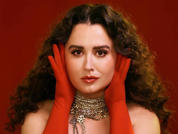 Close-up portrait of a woman with long curly brown hair wearing red gloves and a layered crystal choker necklace against a dark red backdrop.
