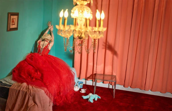 Woman in red gown poses in a room with aqua walls, chandelier, peach curtains, and red carpet, creating a surreal and opulent atmosphere.