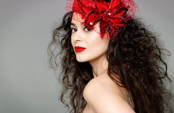 Close-up portrait of a woman with long curly dark hair wearing a red fascinator and bold red lipstick against a grey background.