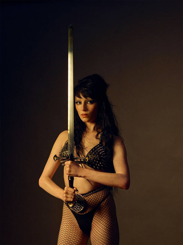 Woman in black bra and fishnet tights holds a large sword in a studio setting with a dark background and soft, focused light.