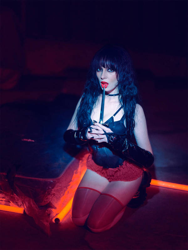 Woman with black hair, wearing a black corset and red lingerie, poses on her knees in a dimly lit room with a dagger in her mouth.