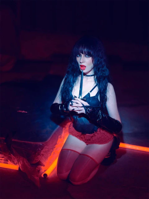 Woman with black hair, wearing a black corset and red lingerie, poses on her knees in a dimly lit room with a dagger in her mouth.