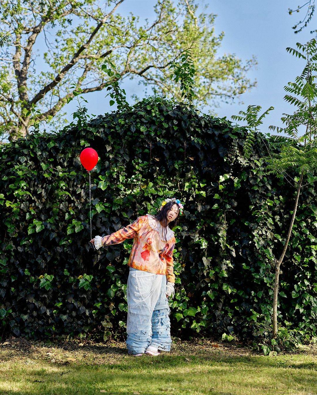 Woman in colorful sweater and white pants standing in a grassy yard with a red balloon in front of a leafy green hedge.