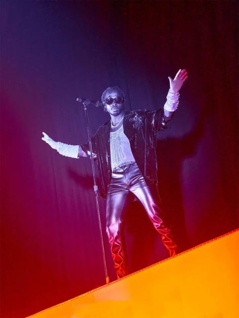Singer on stage with microphone, wearing shiny pants, sheer shirt, and sunglasses in a performance setting with colored lighting.