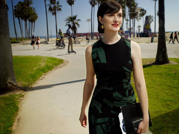 Attractive woman in patterned dress walks on path near beach with palm trees, holding a tablet. Sunny day, outdoor setting.