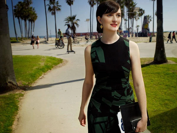 Attractive woman in patterned dress walks on path near beach with palm trees, holding a tablet. Sunny day, outdoor setting.