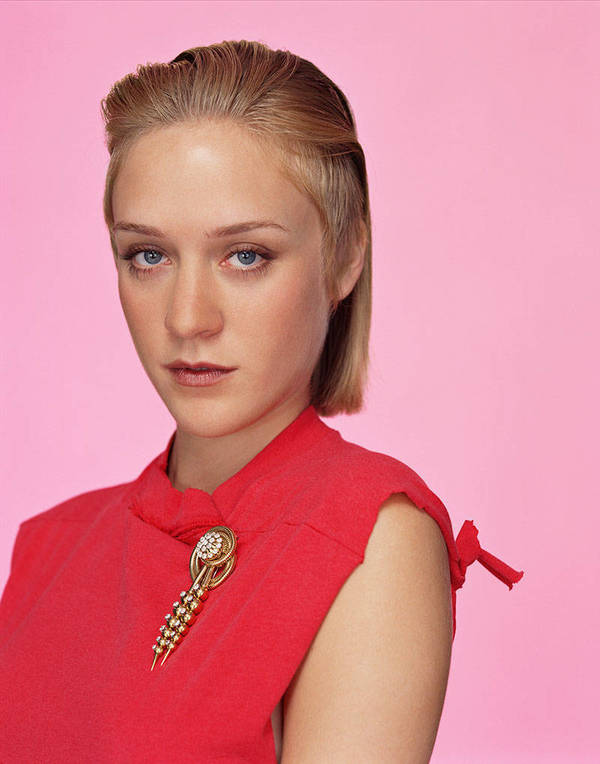 Portrait of a blonde woman with fair skin wearing a red dress with a gold brooch against a pink backdrop in a studio setting.