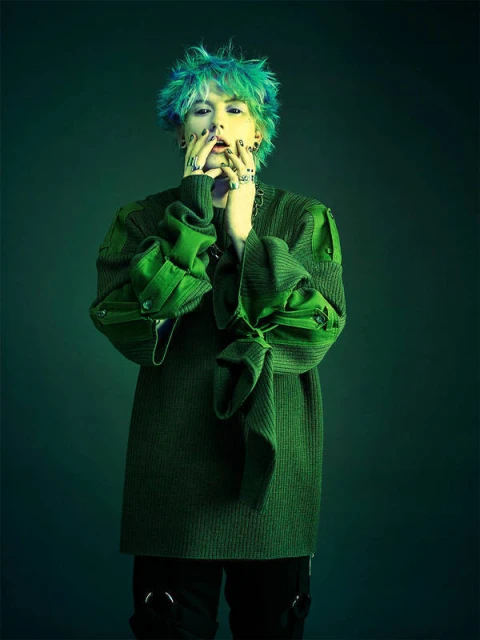Person with green and blue hair wearing a green sweater and black pants poses in front of a dark green backdrop, eye-level shot.