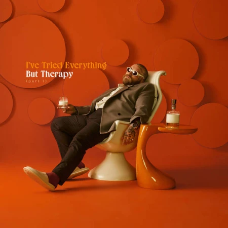 Man wearing sunglasses and a blazer reclining in a white chair against an orange wall with circular reliefs.