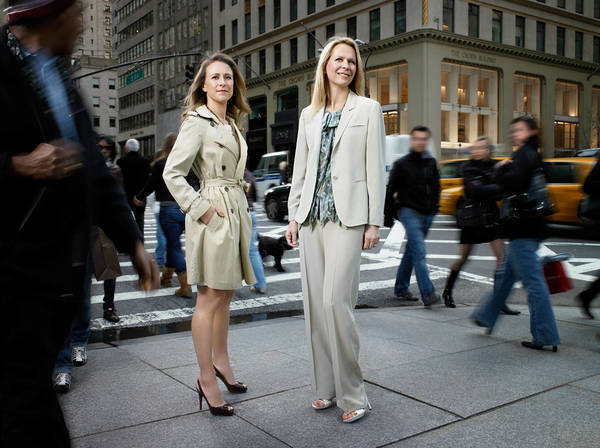 Two adult women stand in a crosswalk on a city street in business attire, surrounded by blurred pedestrians and vehicles in daytime.