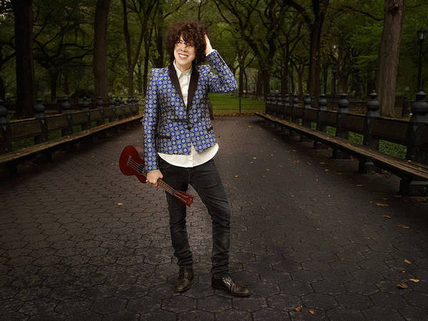 Person stands on a paved path in a park holding a ukulele, wearing a patterned blazer, black jeans, and brown dress shoes.