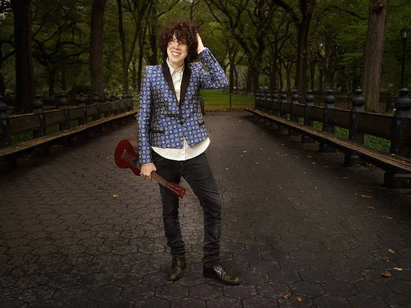 Person stands on a paved path in a park holding a ukulele, wearing a patterned blazer, black jeans, and brown dress shoes.