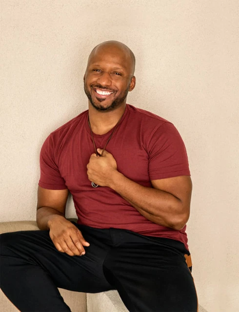 Portrait of smiling African American man with burgundy shirt and black pants sitting in an indoor setting with neutral wall background.
