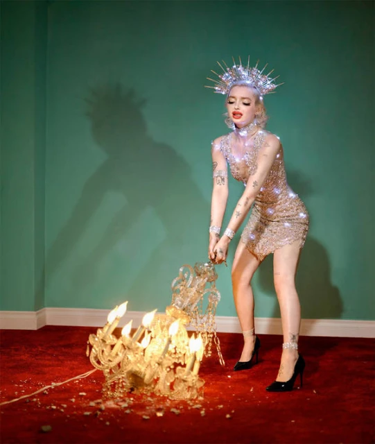 Woman in sparkling dress posing with a crystal chandelier in an indoor scene with a turquoise wall and red floor.