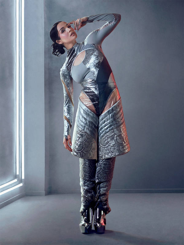 A woman in futuristic grey body suit and silver platforms poses in a grey photo studio with a vertical white neon light.