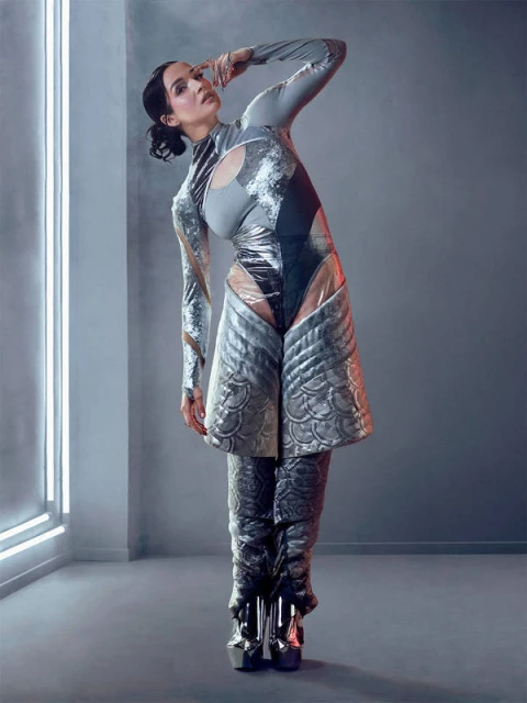 A woman in futuristic grey body suit and silver platforms poses in a grey photo studio with a vertical white neon light.