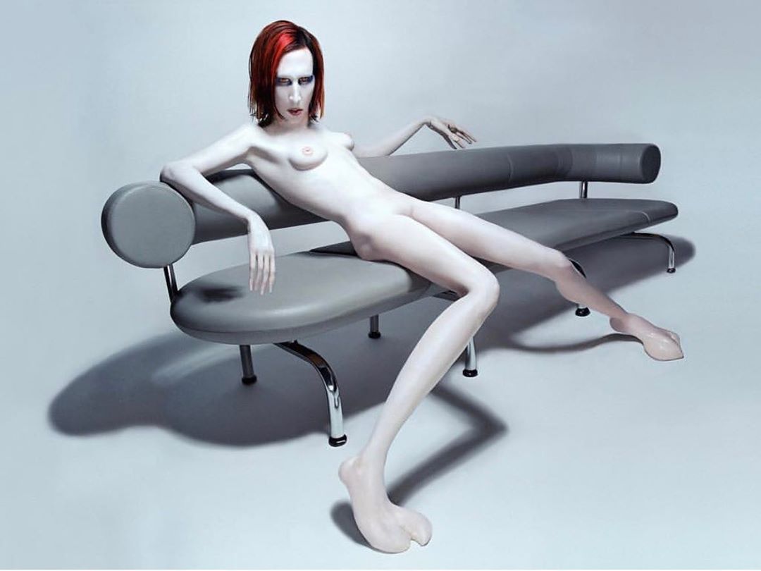Surreal nude adult woman with red hair, posed on a grey modern chaise lounge in a brightly lit, surreal studio with a seamless background.