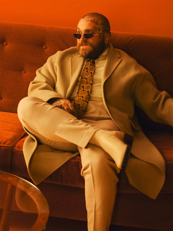 Stylish man with beard and sunglasses lounges on sofa in suit and patterned tie in orange room, exuding retro vibe.