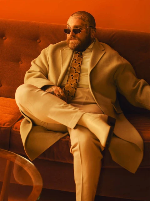 Stylish man with beard and sunglasses lounges on sofa in suit and patterned tie in orange room, exuding retro vibe.