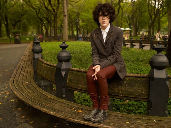 A young adult sits on a park bench wearing a grey blazer and rust-colored jeans, looking thoughtfully at the camera in a verdant park.