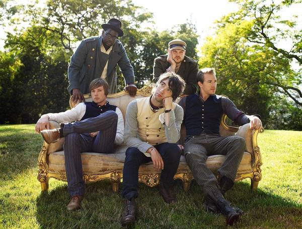 Five men sit outside on an ornate couch, casually posed in dress clothes, surrounded by greenery and natural light.