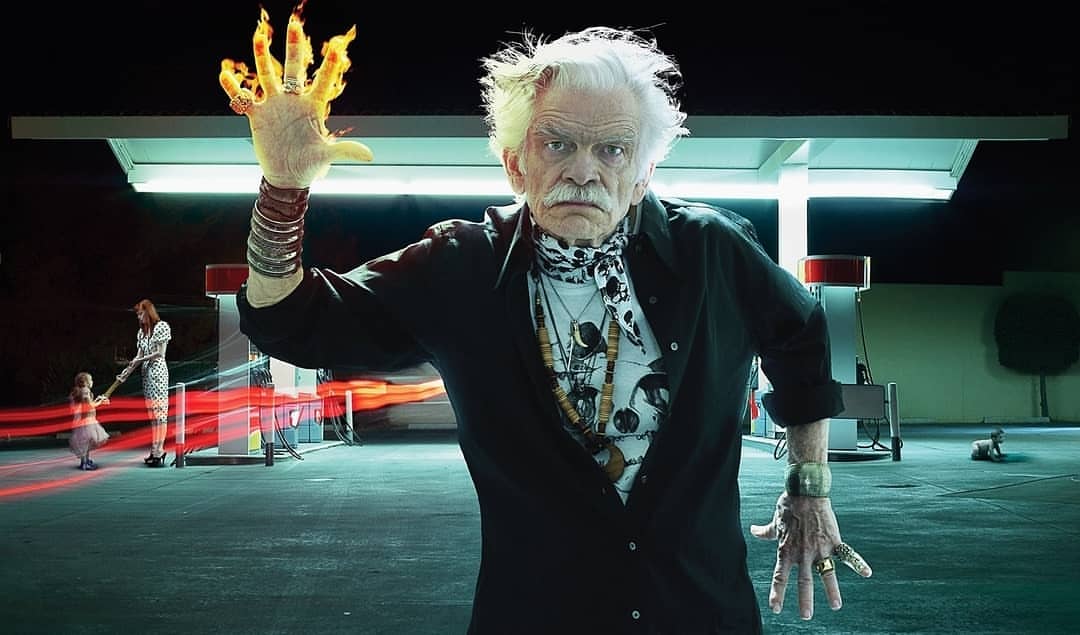 Elderly man with a fiery hand standing at a gas station at night, with a woman and child in the background and a dog.
