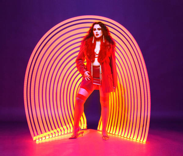 Fashionable woman in red coat and stocking, stands center against purple background and lit tunnel, posed in front of studio lighting.
