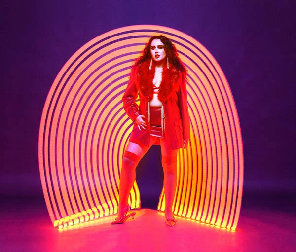 Fashionable woman in red coat and stocking, stands center against purple background and lit tunnel, posed in front of studio lighting.