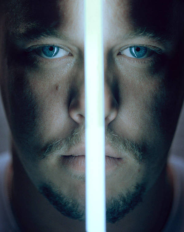 Close-up portrait of an adult male with light stubble, bright blue eyes, and a bright linear light bisecting his face vertically.