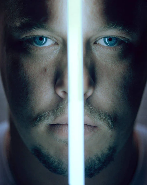 Close-up portrait of an adult male with light stubble, bright blue eyes, and a bright linear light bisecting his face vertically.