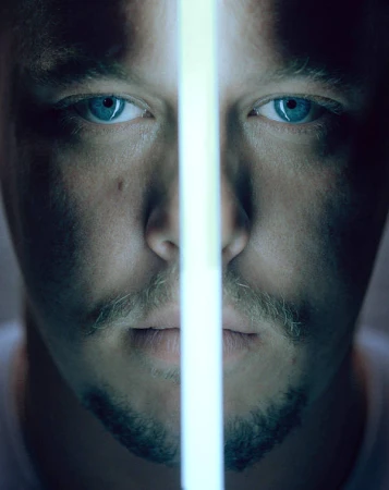 Close-up portrait of an adult male with light stubble, bright blue eyes, and a bright linear light bisecting his face vertically.