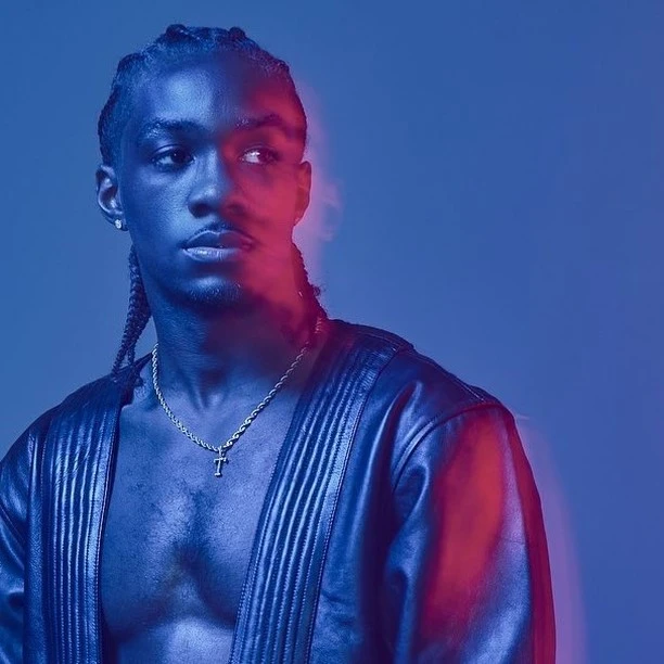 Studio portrait of a dark-skinned man in an open leather jacket against a blue background with red rim lighting and wearing braids.