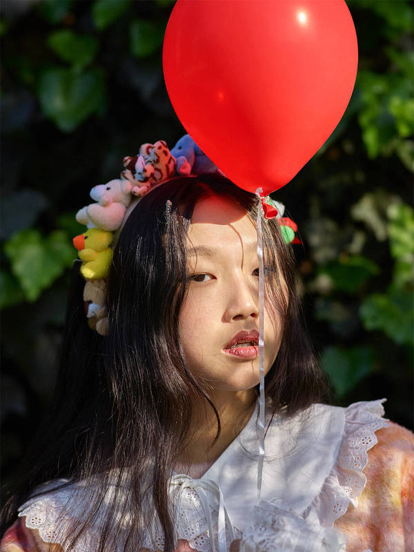 Young woman poses outdoors in playful portrait, adorned with a stuffed-animal headband and holding a red balloon on a sunny day.