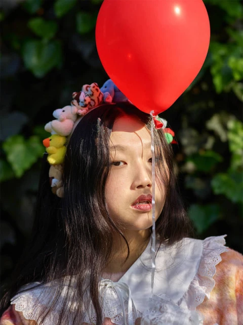 Young woman poses outdoors in playful portrait, adorned with a stuffed-animal headband and holding a red balloon on a sunny day.
