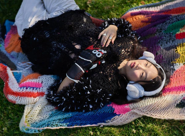 Adult woman lying down on a colorful knitted blanket wearing a black textured top with white trim and white furry earmuffs outdoors in grass.