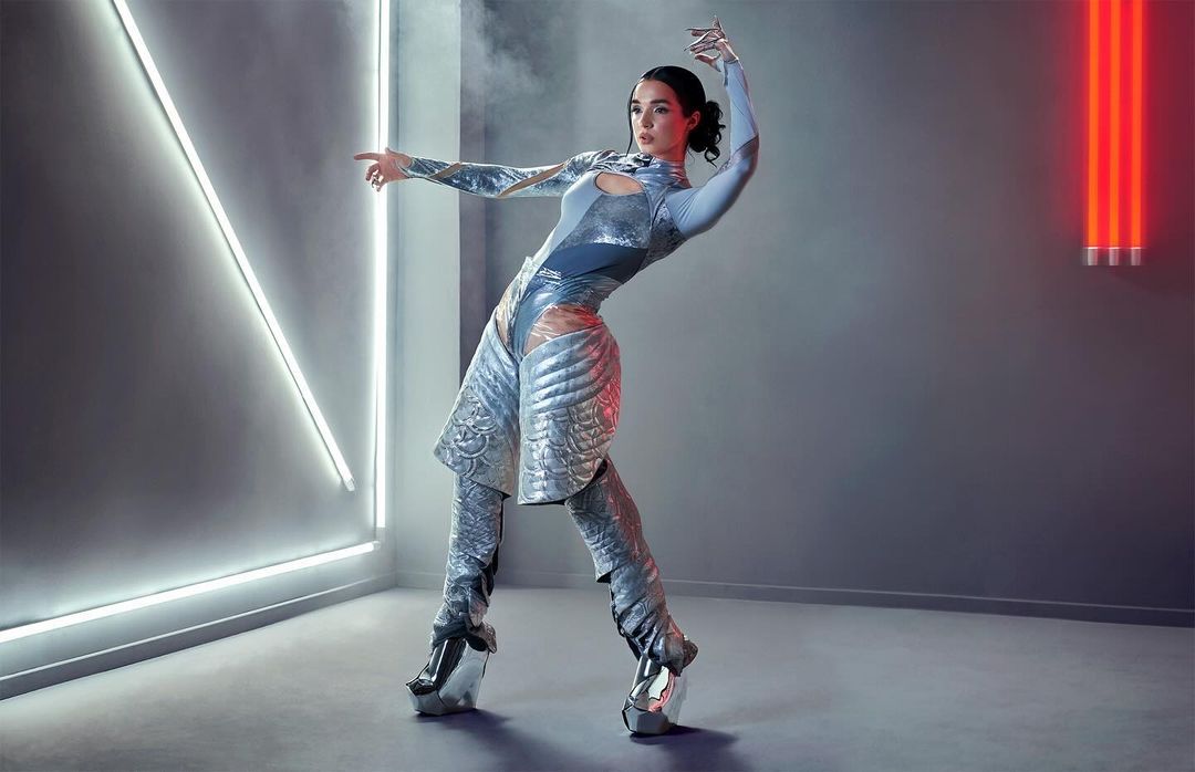 A woman in a futuristic bodysuit and platform shoes poses in a studio with white and red neon lights, creating a dynamic and visually striking image.