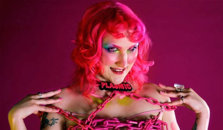 Adult female with pink hair, colourful makeup, and chained in pink stands in front of solid pink backdrop in a studio-lit setting.