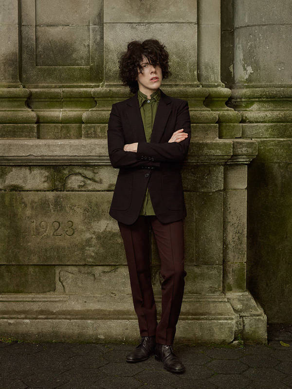 Non-binary individual in a dark blazer, light green shirt, and dark pants stands against a stone wall with carved details.