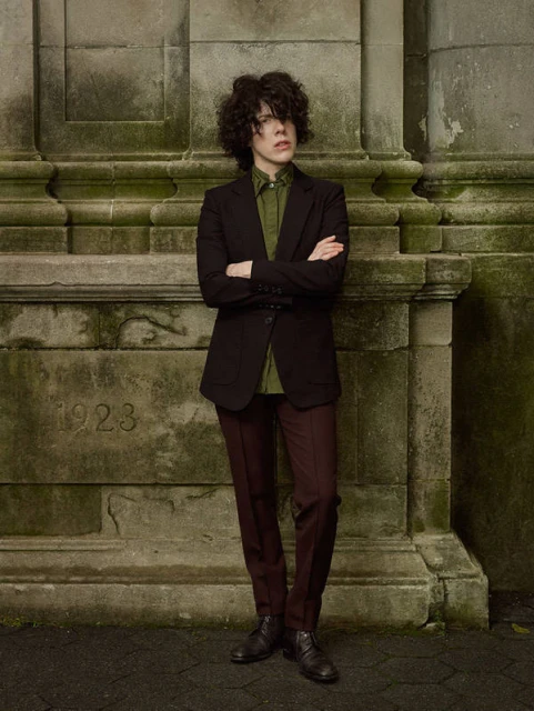 Non-binary individual in a dark blazer, light green shirt, and dark pants stands against a stone wall with carved details.