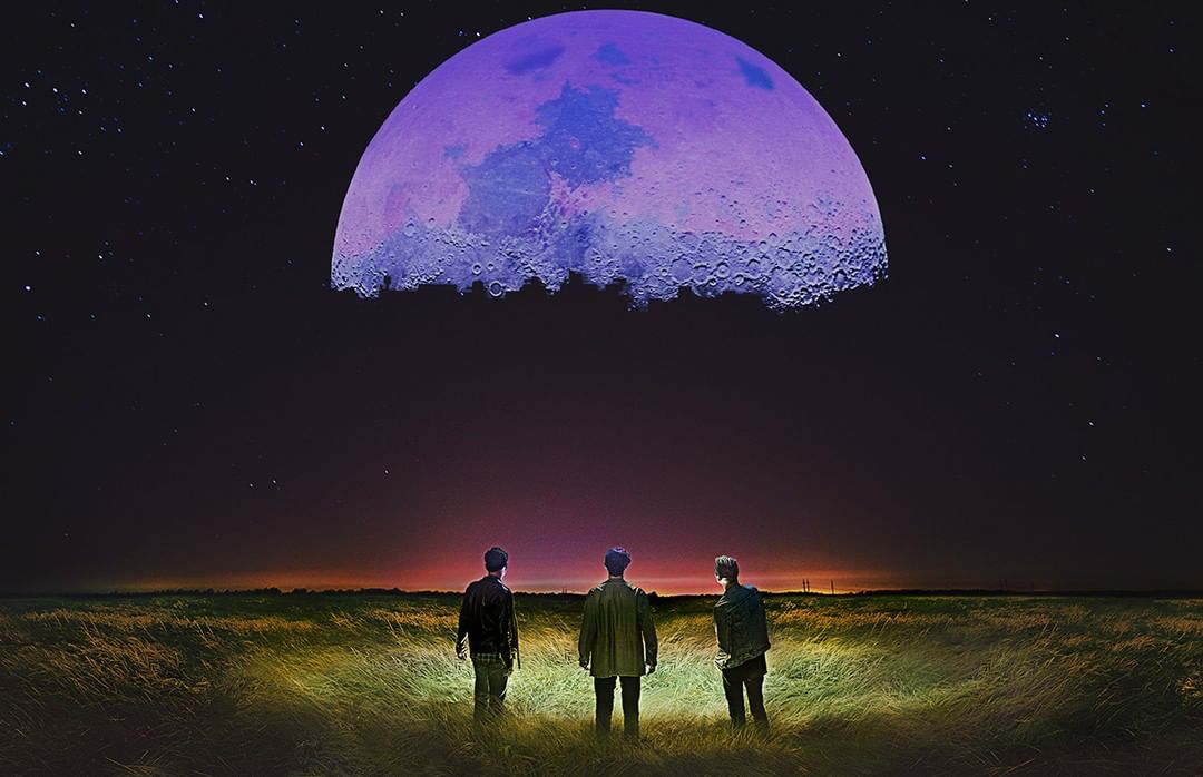Three men stand in a grassy field under a giant purple moon at night, with stars visible in the dark sky.