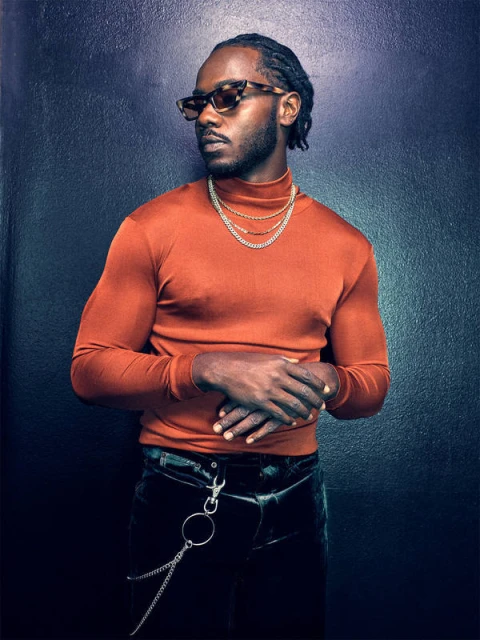 Portrait of a Black male wearing orange turtleneck, sunglasses, and chains against dark blue backdrop. Fashion, style, and portraiture.