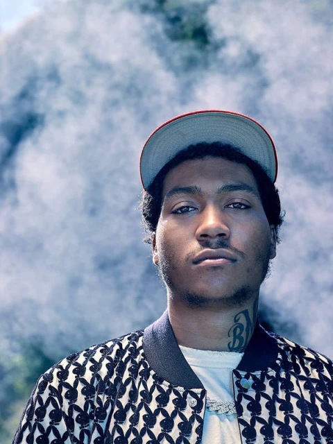Portrait of a young adult male wearing a baseball cap and patterned jacket, gazing directly at the camera with a smoky background.