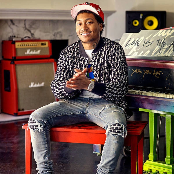Young man sits on a red stool indoors, wearing a patterned jacket and ripped jeans, with a colorful piano and Marshall amps in the background.