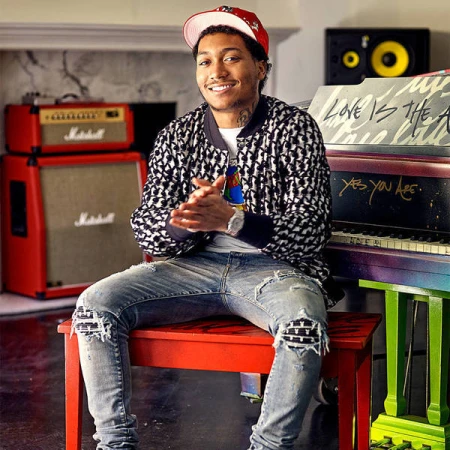 Young man sits on a red stool indoors, wearing a patterned jacket and ripped jeans, with a colorful piano and Marshall amps in the background.
