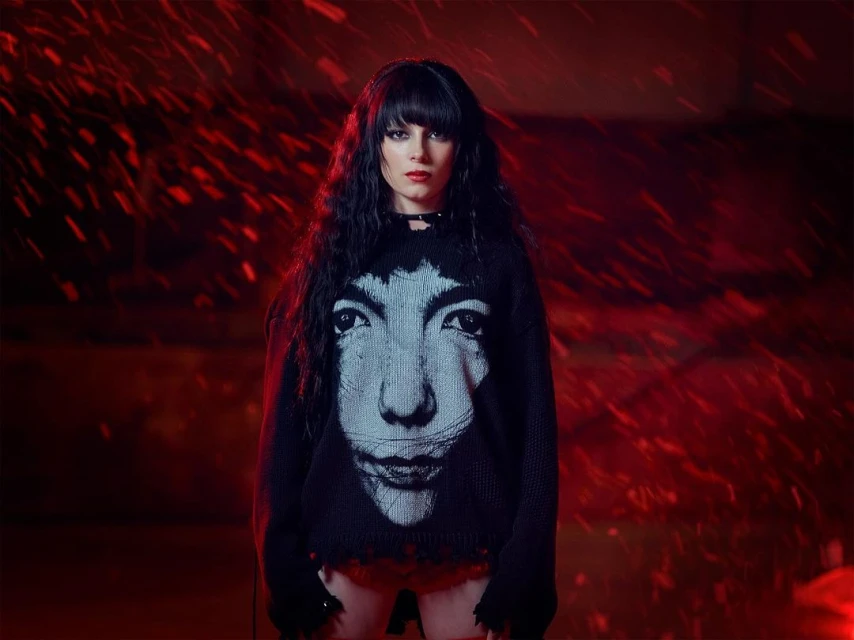 A woman with dark hair stands in front of a red background, wearing a black sweater with a portrait on the front.