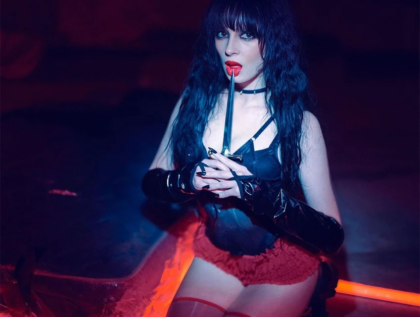 A woman with dark hair and bangs wearing a black corset, red bottoms and gloves poses indoors with a dagger in her mouth.
