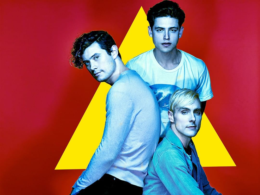 Promotional shot of band LANY, featuring three young adult males, wearing casual clothing, against a vibrant red and yellow geometric backdrop.